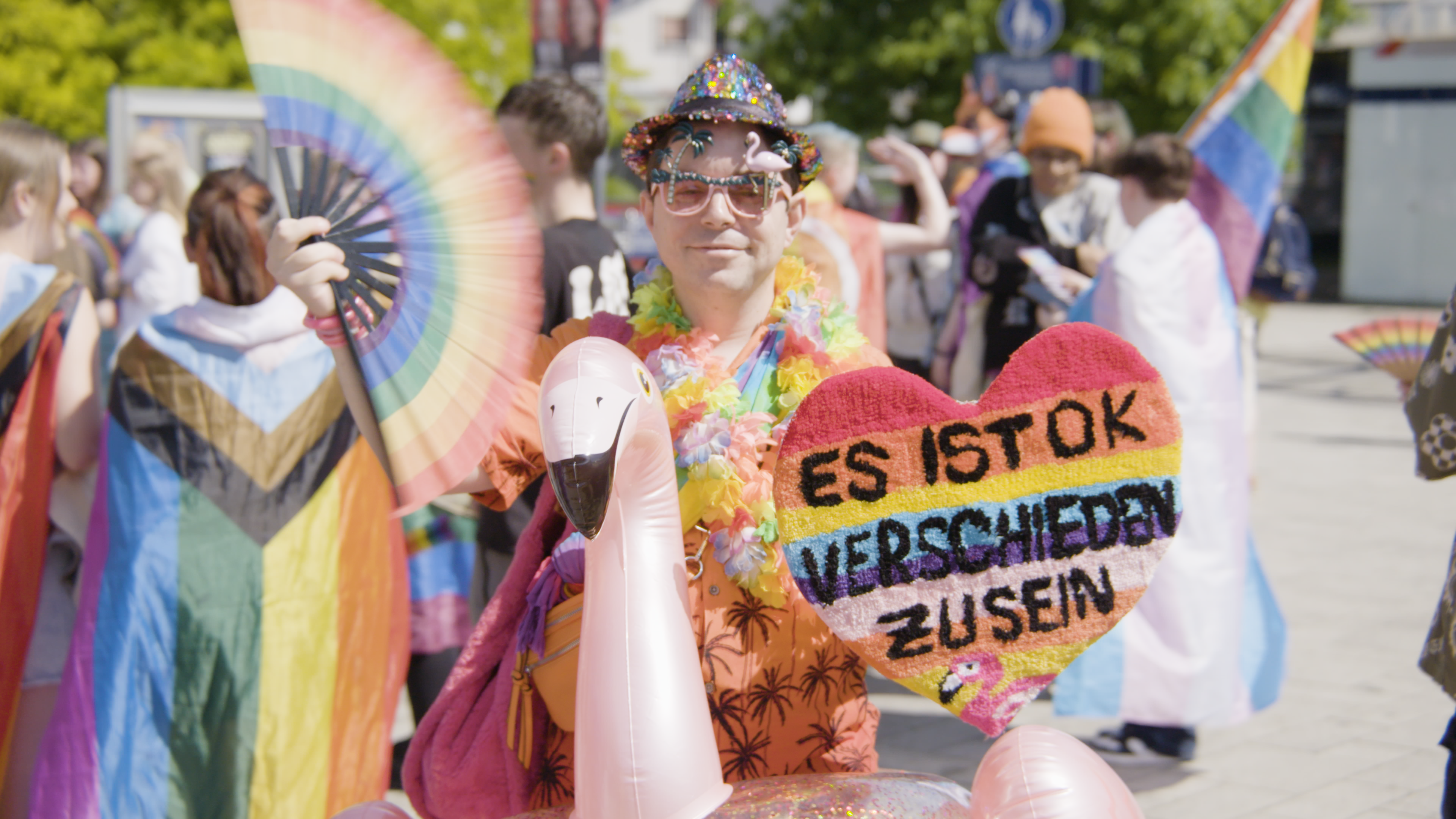 Eine in regenbogenfarben bunt gekleidete Person hält ein Demonstrationsschild in der Hand, auf dem "Es ist ok verschieden zu sein" steht.