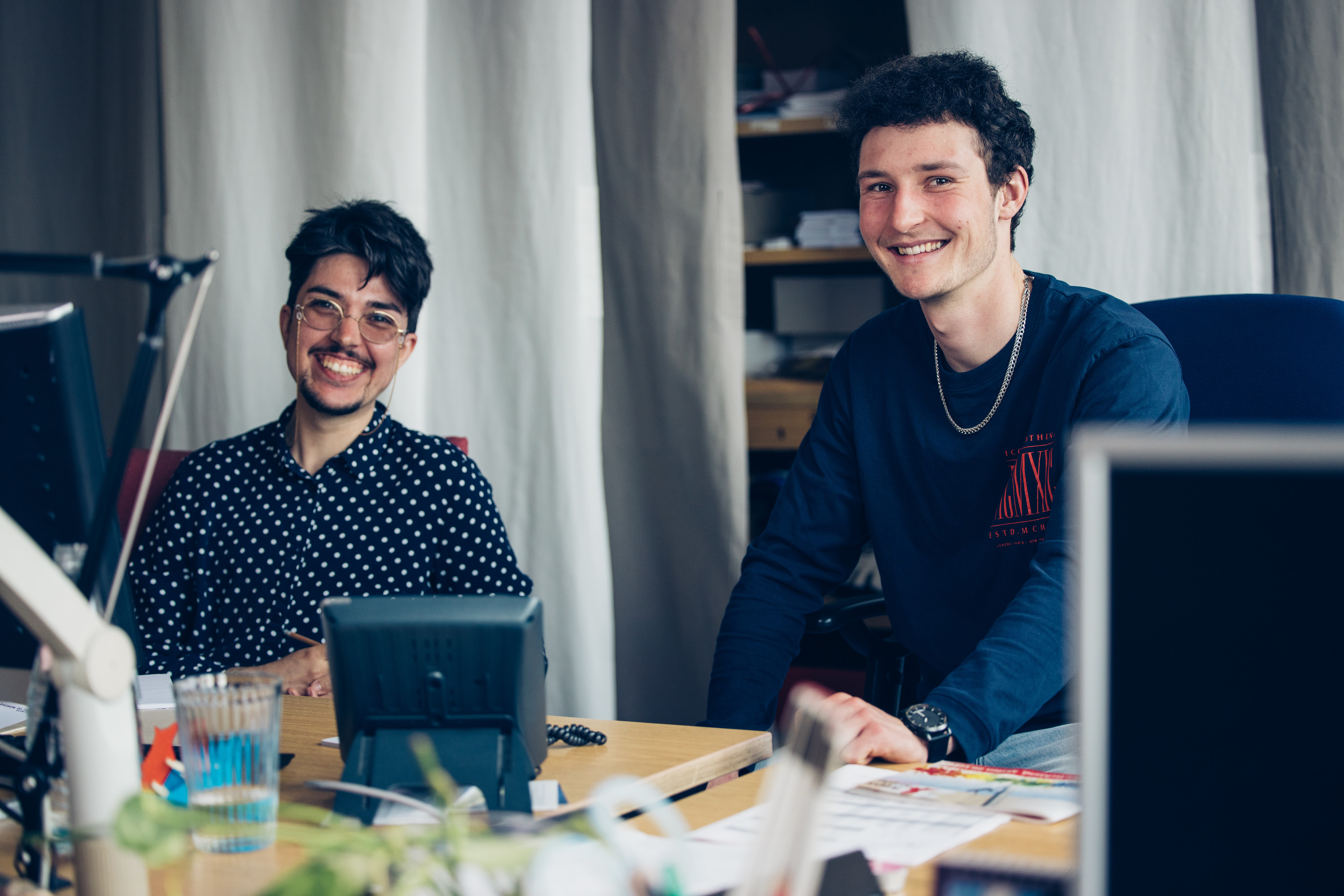 Lien Woywood (links) und Mats K. (rechts) sitzen an einem Schreibtisch in einem Büro und schauen lächelnd in die Kamera.