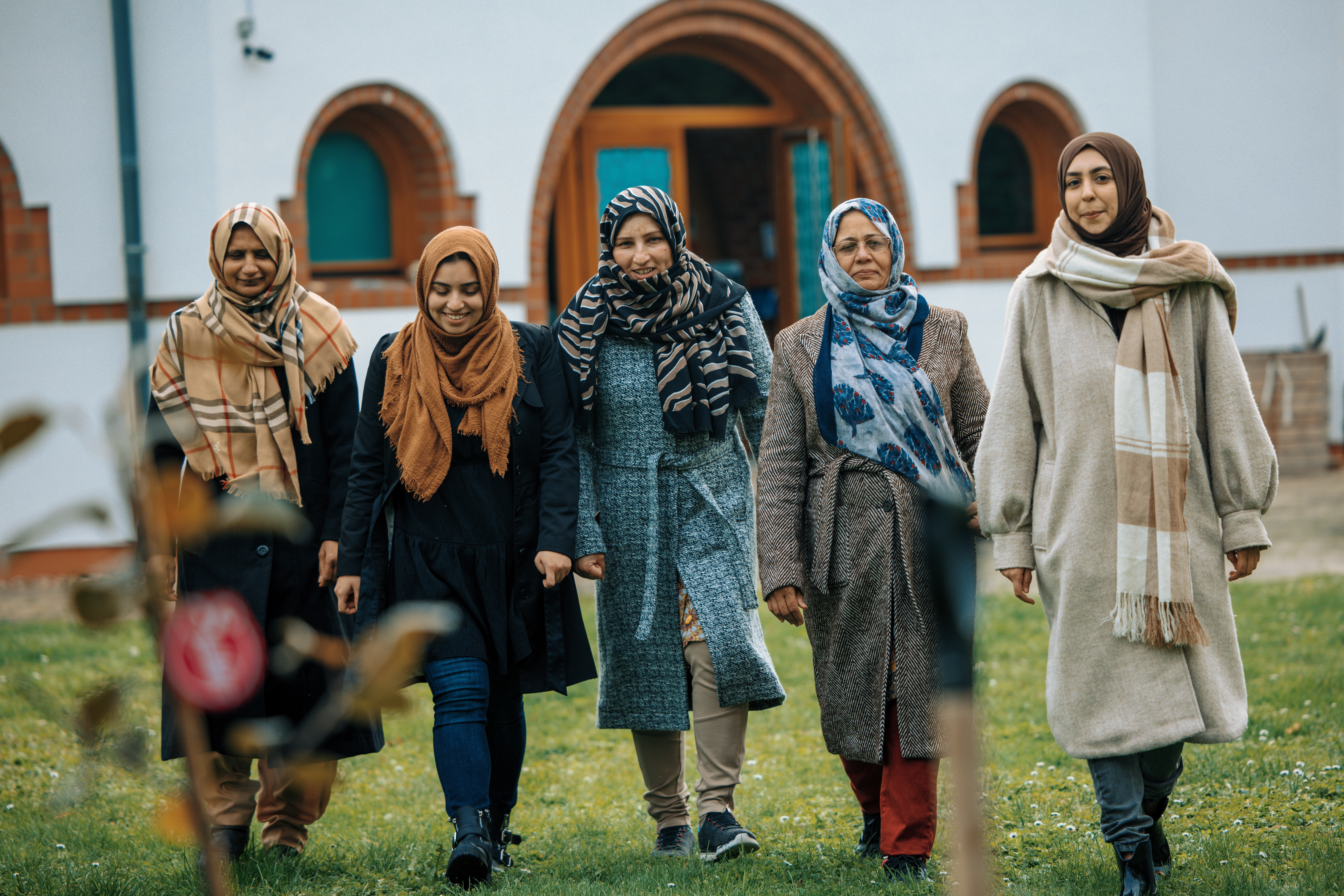 Fünf Frauen laufen lächelnd nebeneinander über eine Wiese vor der Moschee