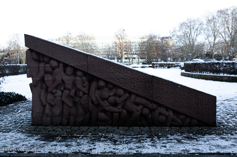 Auf einer schneebedeckten Wiese steht ein rot-brauner Sandsteinblock mit stilisierten Figuren im Hochrelief.