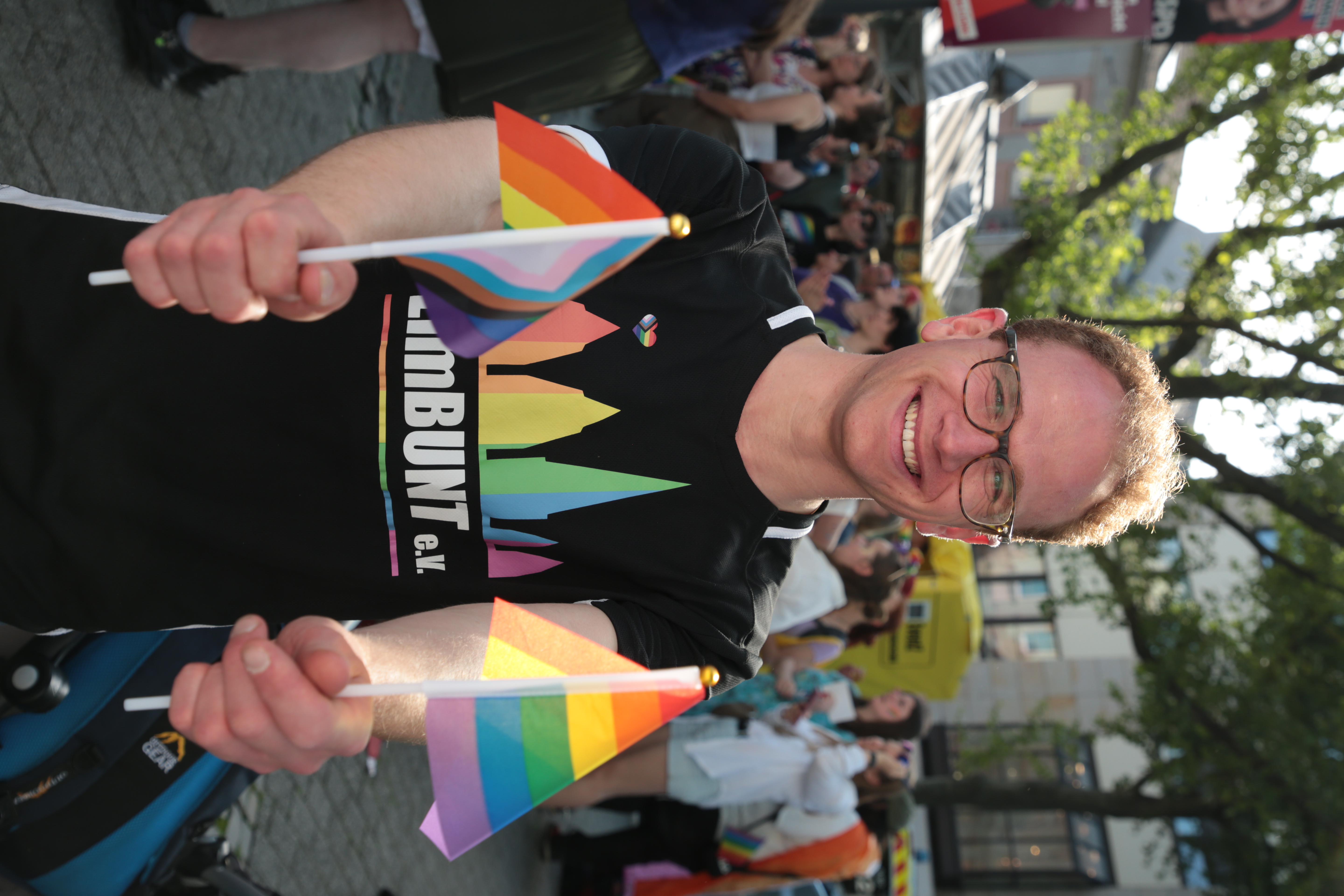 Eine Person mit Brille hält eine Regenbogenflagge in der einen und eine Progress Flagge in der anderen Hand und blickt glücklich in die Kamera.