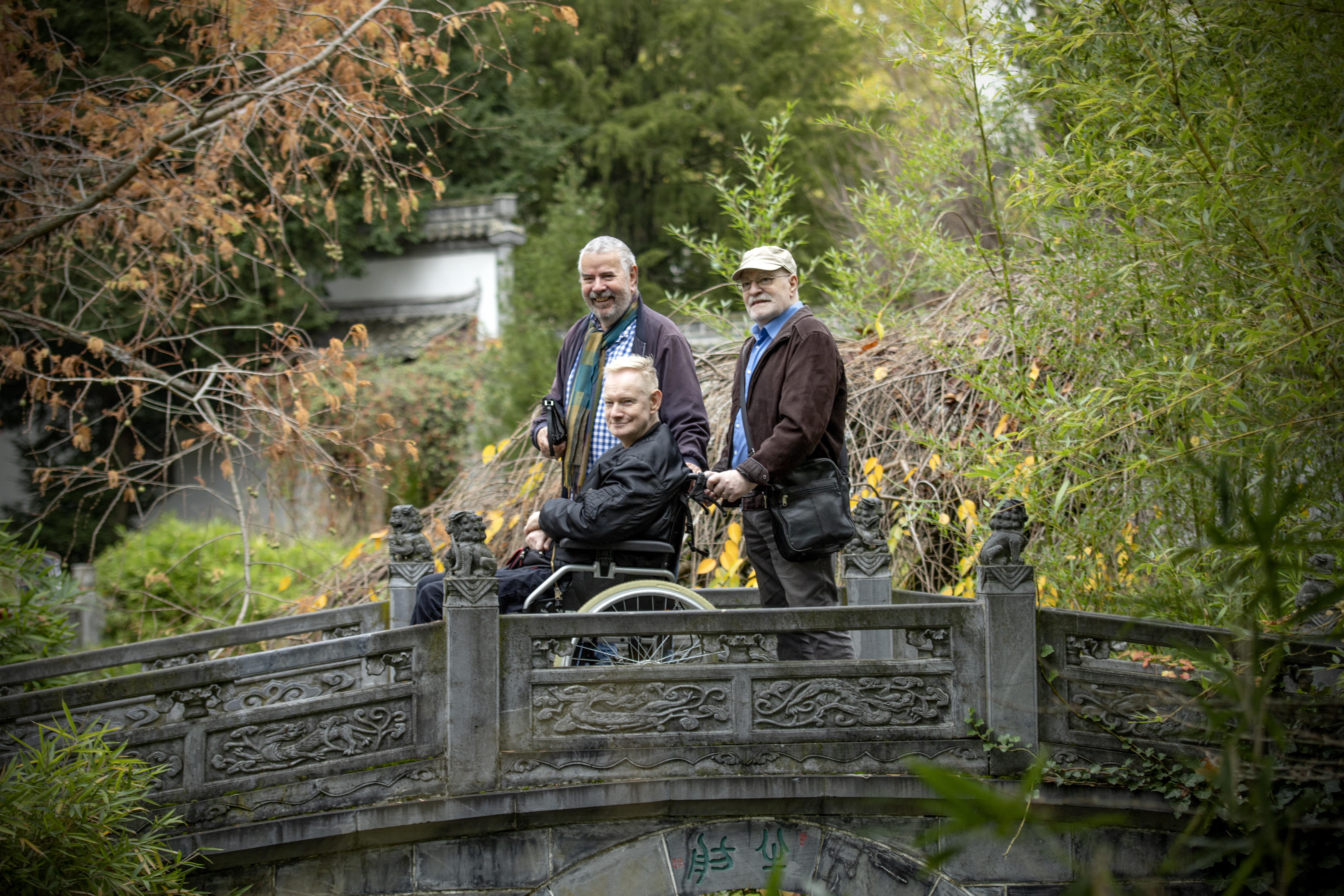 Antonio, Bernd und Detlef sind auf einer kleinen Brücke in einem Garten. Antonio hält Bernds Rollstuhl. Alle drei blicken über ihre linke Schulter in die Kamera
