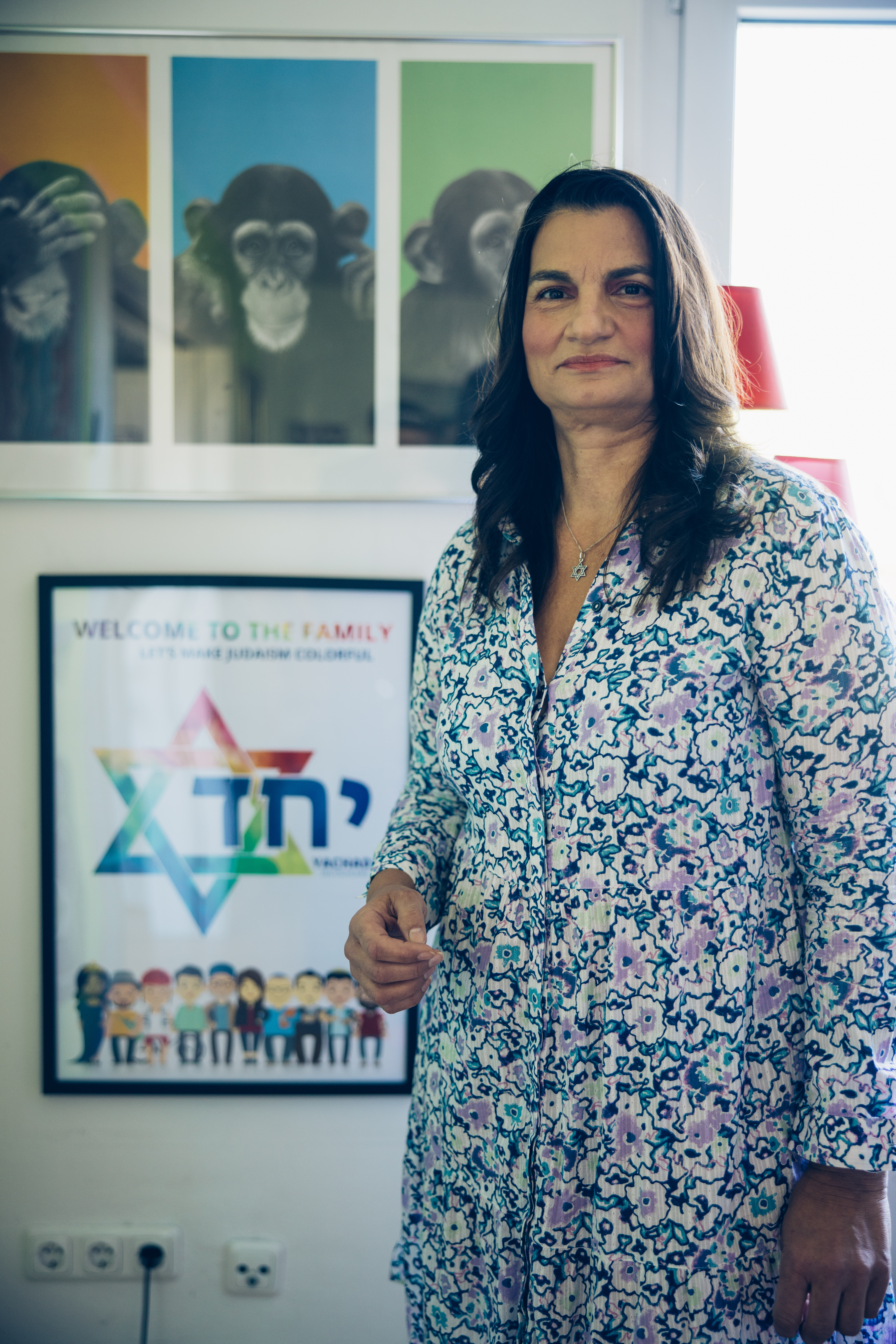 Eine Frau mit langen schwarzen Haaren und buntem Kleid steht vor einem regebogenbunten Plakat des queer-jüdischen Vereins Yachad e.V.