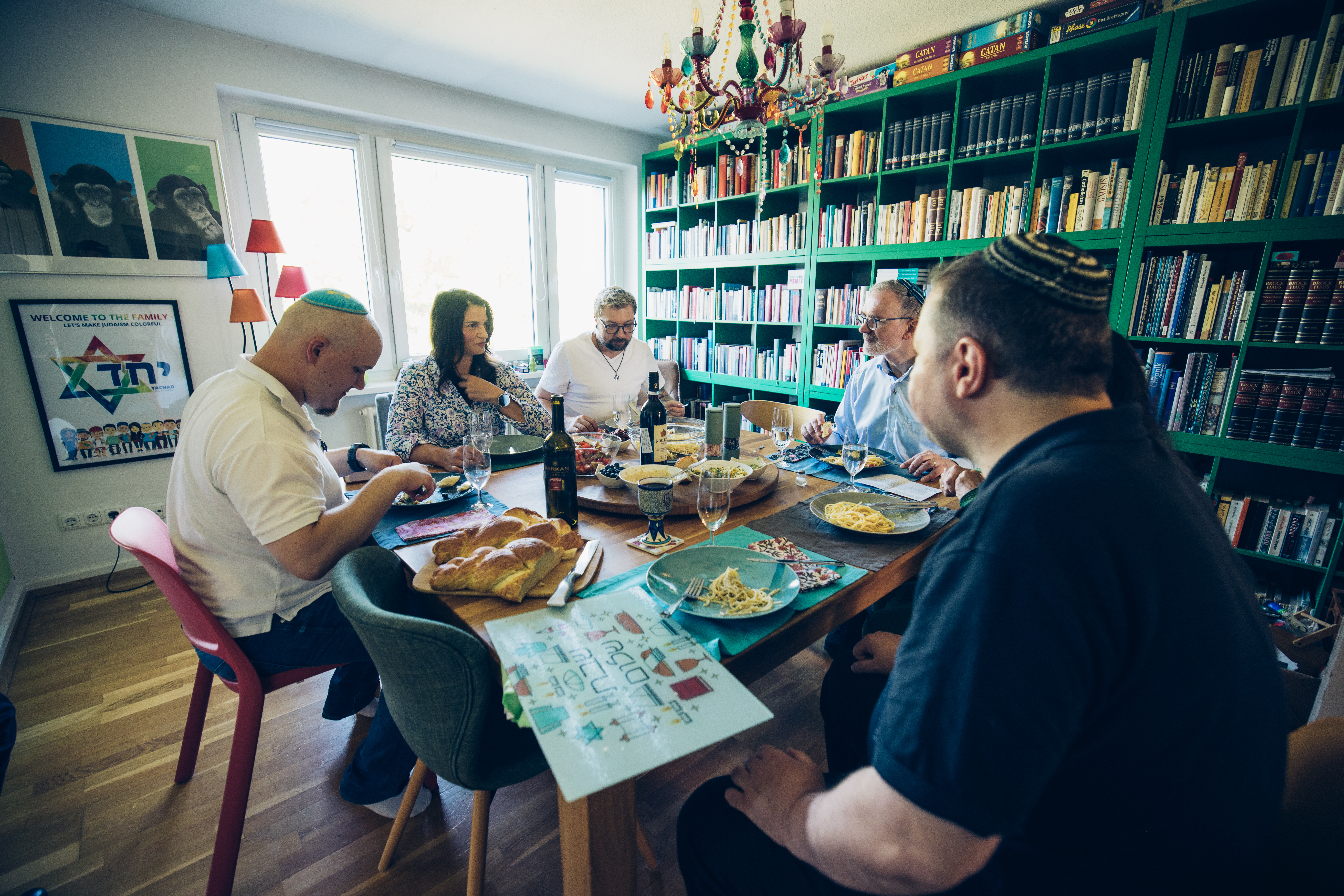 Fünf Menschen sitzen an einem großen gedeckten Esstisch und unterhalten sich. Vier Personen tragen Kippas auf dem Kopf. Auf dem Tisch stehen tradionell jüdische Speisen.